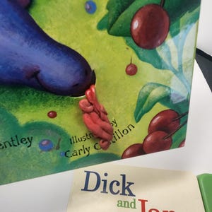 Puede incluir: Un libro infantil con una portada verde que presenta ilustraciones de cerezas y una criatura morada. El t&iacute;tulo del libro es parcialmente visible, junto con el texto "Ilustrado por Carly Cillon". Tambi&eacute;n se ve otro libro titulado "Dick and Jane".