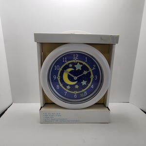 May include: A round 10-inch diameter wall clock with a white plastic frame and a dark blue face featuring a yellow crescent moon and light green stars. The clock has silver hands, white numbers, and is packaged in a cardboard box with text.