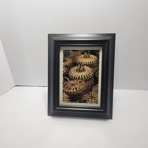 May include: A framed print featuring a close-up of several woven baskets. The baskets are primarily tan and brown, with intricate patterns. The frame is a simple, rectangular black frame, adding a touch of contrast to the natural tones of the artwork.