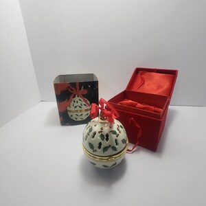 Könnte beinhalten: Eine weiße Keramik-Weihnachtskugel mit Stechpalmen-Beeren-Design, goldfarbenem Rand und rotem Band. Die Kugel befindet sich neben einer roten Samtbox und einer passenden Geschenkbox mit ähnlichem Design.