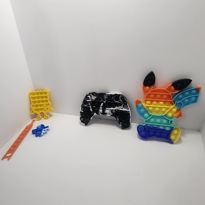 May include: A set of four colorful pop-it fidget toys. The toys are shaped like a video game controller, a cartoon character, and two simple geometric shapes. The toys are made of silicone and have a variety of colors, including black, white, orange, blue, and yellow.
