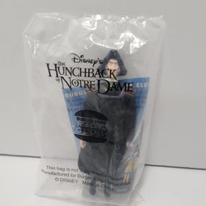 May include: A plastic action figure of a character from Disney's "The Hunchback of Notre Dame" movie, inside a clear plastic bag. The bag has the movie title and Burger King Kids Club branding.