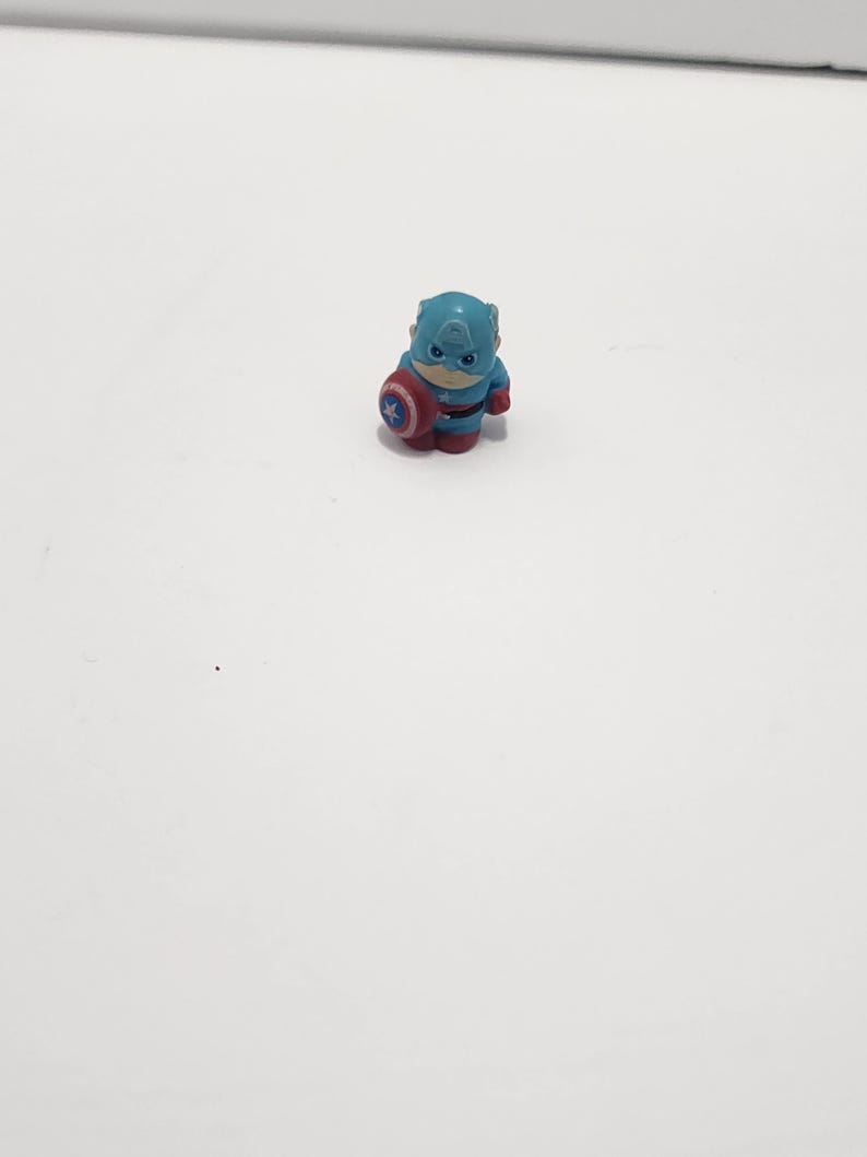 May include: A small blue and red plastic figurine of Captain America holding his shield. The figurine is about 2 inches tall.