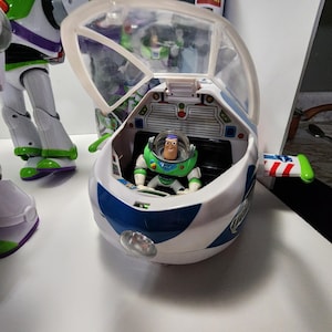 May include: A white and blue toy spaceship with a clear plastic canopy. A green and purple toy astronaut figure is sitting in the cockpit. The spaceship has a red, white, and blue design on the side and a light on the front. The toy is from the Disney Pixar Toy Story franchise.