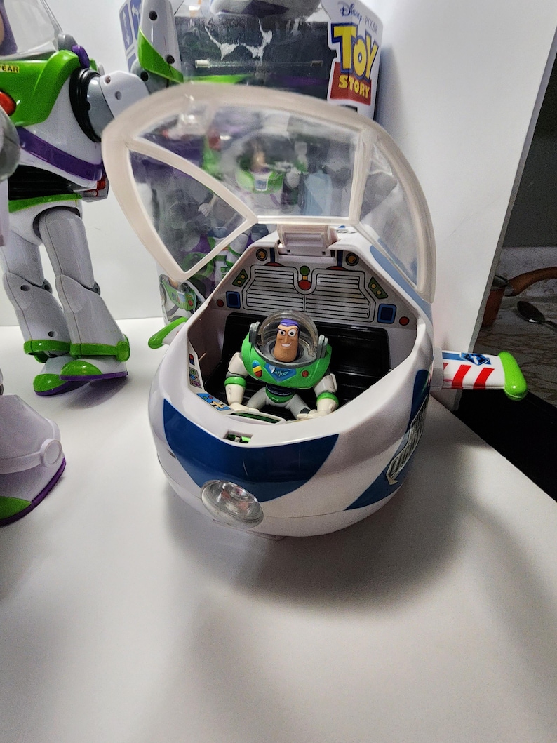 May include: A white and blue toy spaceship with a clear plastic canopy. A green and purple toy astronaut figure is sitting in the cockpit. The spaceship has a red, white, and blue design on the side and a light on the front. The toy is from the Disney Pixar Toy Story franchise.