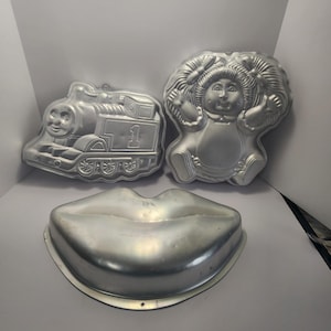 Puede incluir: Tres moldes para pasteles de aluminio vintage en varias formas. Uno es un tren Thomas the Tank Engine, otro es un Cabbage Patch Kid y el tercero es un par de labios. Ideales para hornear pasteles temáticos.