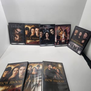 Puede incluir: Una colección de ocho DVD de la serie Twilight, incluyendo Twilight, New Moon, Eclipse, Breaking Dawn Part 1 y Breaking Dawn Part 2. Las carátulas muestran retratos de personajes y títulos en texto blanco y dorado sobre fondos oscuros.