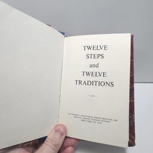 Könnte beinhalten: Aufgeschlagenes Buch mit dem Titel "TWELVE STEPS and TWELVE TRADITIONS" in schwarzer Schrift. Der Einband des Buches hat ein rotes und goldenes Blumenmuster. Der Text "ALCOHOLICS ANONYMOUS WORLD SERVICES, INC." ist ebenfalls sichtbar.