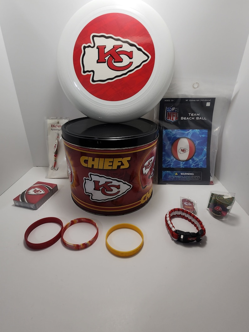 May include: A collection of Kansas City Chiefs fan merchandise, including a white and red frisbee, a Chiefs-branded tin, a beach ball, bracelets, and a deck of cards. The items feature the team's logo in red and gold.