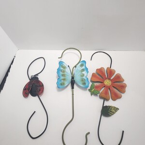 Peut inclure: Trois crochets de jardin en métal avec des accents décoratifs. Les crochets sont en forme de coccinelle, de papillon et de fleur. La coccinelle est rouge avec des points noirs, le papillon est bleu avec des marques noires et la fleur est orange avec un centre jaune.