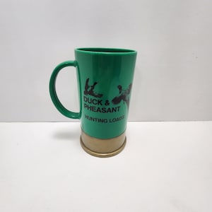 Puede incluir: Taza de cerámica verde con forma de cartucho de escopeta con una imagen en blanco y negro de un pato y una perdiz y el texto "Duck & Pheasant Hunting Loads".