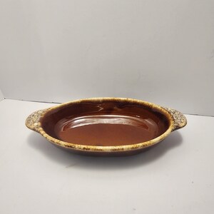 Pode incluir: Assadeira oval de cerâmica marrom com duas alças. A assadeira tem um interior brilhante e uma borda texturizada e salpicada. As alças também são texturizadas. A assadeira é provavelmente para assar e servir.