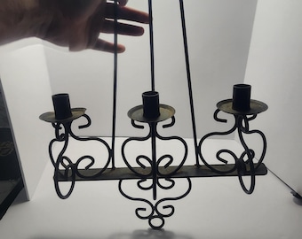 Puede incluir: Candelabro de hierro forjado negro con tres portavelas y adornos decorativos. El candelabro está suspendido por tres varillas verticales. El diseño presenta elementos en forma de corazón y un adorno central.