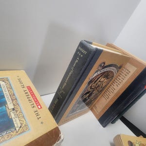 May include: A collection of hardcover books from the "A Series of Unfortunate Events" series by Lemony Snicket. The books have tan and blue covers with illustrations and text. One book is titled "The Slippery Slope".