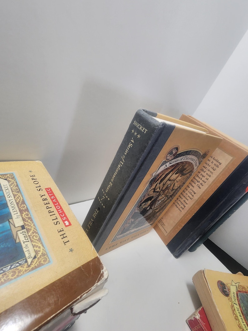 May include: A collection of hardcover books from the "A Series of Unfortunate Events" series by Lemony Snicket. The books have tan and blue covers with illustrations and text. One book is titled "The Slippery Slope".
