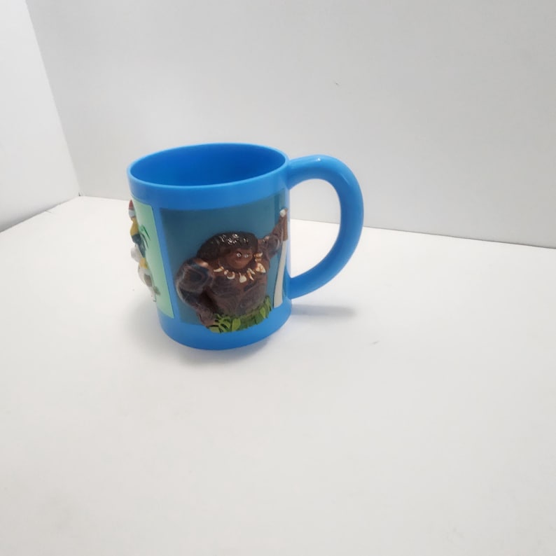 May include: A blue ceramic mug with a curved handle. The mug features a 3D relief of a cartoon character on the side. The character is brown with a grass skirt and a necklace. The mug is on a white surface.