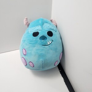 May include: A light blue plush toy of the character Sulley from Monsters, Inc. The toy features darker blue markings, two small horns, and purple spots. It has a smiling face with black eyes and a single tooth.