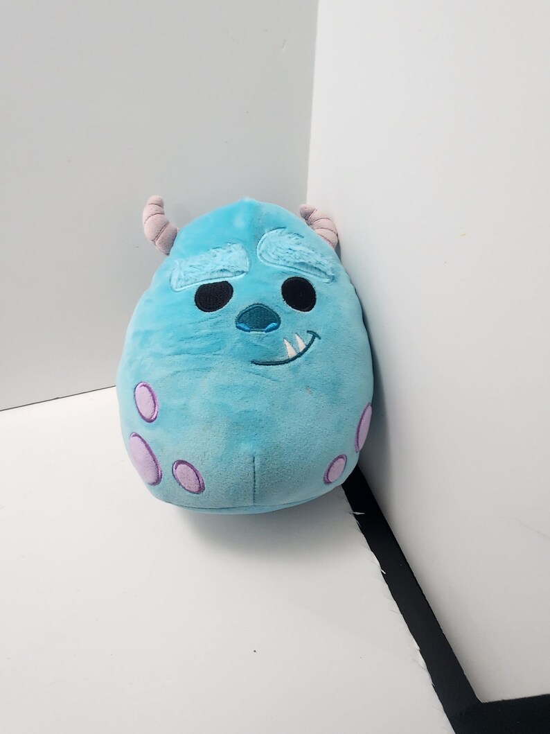 May include: A light blue plush toy of the character Sulley from Monsters, Inc. The toy features darker blue markings, two small horns, and purple spots. It has a smiling face with black eyes and a single tooth.