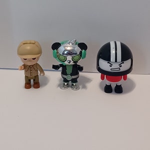 Puede incluir: Tres peque&ntilde;as figuras de pl&aacute;stico. Una figura marr&oacute;n y beige con un sombrero y una camisa a cuadros. Una figura negra y verde con una cara de panda y detalles plateados. Una figura negra y blanca con un casco y una camisa roja.