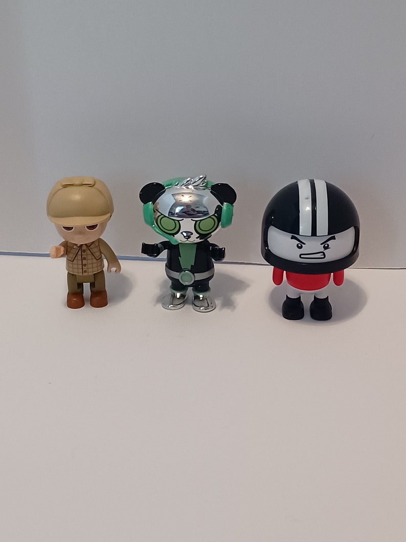 Puede incluir: Tres peque&ntilde;as figuras de pl&aacute;stico. Una figura marr&oacute;n y beige con un sombrero y una camisa a cuadros. Una figura negra y verde con una cara de panda y detalles plateados. Una figura negra y blanca con un casco y una camisa roja.