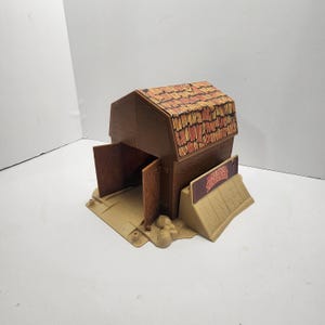 May include: A brown plastic toy barn with a tan base and a red and yellow roof. The barn has two open doors and a sign that reads "Ranch Hand".
