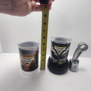 May include: Two clear plastic Monster Jam cups, one measuring approximately 4 inches tall. One cup sits on a black tire-shaped base. A silver metal piston and connecting rod are also present. The cups feature the Monster Jam logo.