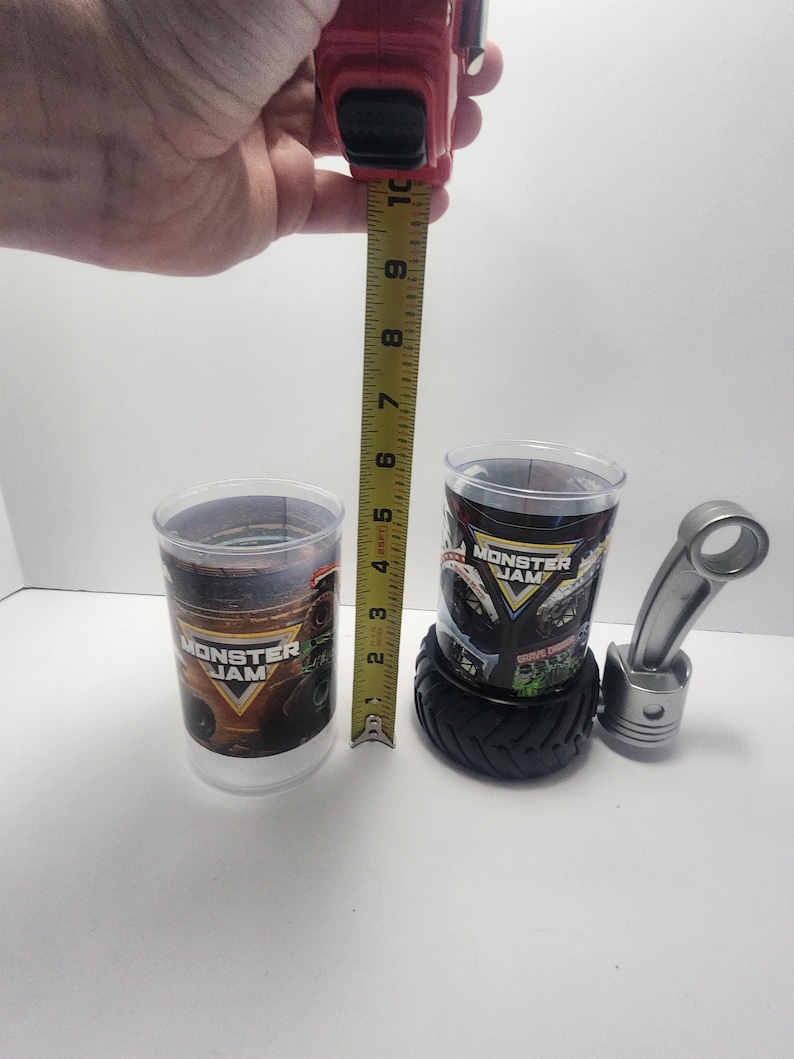 May include: Two clear plastic Monster Jam cups, one measuring approximately 4 inches tall. One cup sits on a black tire-shaped base. A silver metal piston and connecting rod are also present. The cups feature the Monster Jam logo.