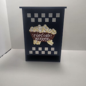May include: A navy blue popcorn keeper with a shelf. The front features a brown bowl filled with popcorn and the words "Popcorn Keeper" in white. White and navy checkered patterns are at the top and bottom.