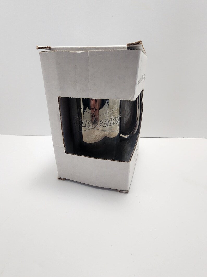 Puede incluir: Una caja de cart&oacute;n blanca con una taza de acero inoxidable plateada en su interior. La taza tiene el logotipo de Budweiser grabado en el lateral.