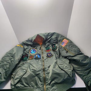 May include: A green bomber jacket with multiple patches and an American flag patch. The jacket has a gold zipper and a green ribbed collar and cuffs. The interior lining is orange.