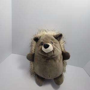 May include: A soft toy hedgehog, featuring a brown body and a spiky, faux-fur back. It has small, rounded ears, a black nose, and a friendly expression. The toy is designed for children.