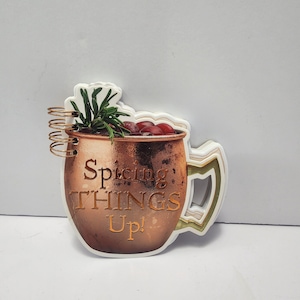 Könnte beinhalten: Kupferfarbene, becherförmige Dekoration mit dem Text "Spicing THINGS Up!". Der Becher ist mit Zweigen grüner Kräuter und roten Beeren gefüllt. Der Griff ist weiß und grün. Die Dekoration hat einen weißen Rand und eine goldene Spirale.