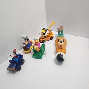 Op de afbeelding: Een verzameling Disney-personagespeelgoed, waaronder Mickey Mouse, Goofy en Donald Duck. Het speelgoed staat in verschillende voertuigen en poses, met heldere kleuren zoals oranje, blauw en groen. Het speelgoed is gemaakt van plastic.