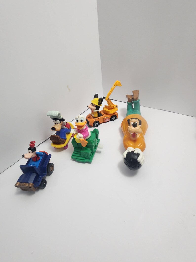 Op de afbeelding: Een verzameling Disney-personagespeelgoed, waaronder Mickey Mouse, Goofy en Donald Duck. Het speelgoed staat in verschillende voertuigen en poses, met heldere kleuren zoals oranje, blauw en groen. Het speelgoed is gemaakt van plastic.