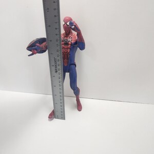 May include: A Spider-Man action figure, approximately 15 cm tall, in a classic red and blue costume. The figure is posed with one arm raised and a web-like accessory. The figure is standing on a white surface.