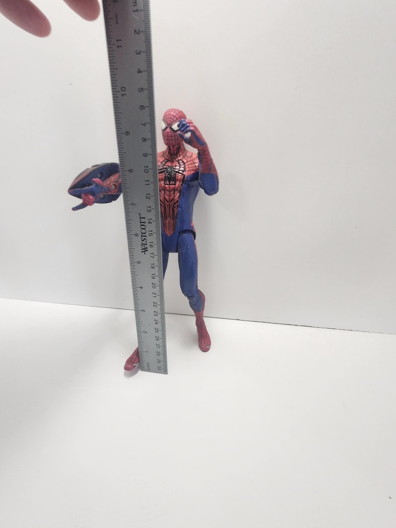 May include: A Spider-Man action figure, approximately 15 cm tall, in a classic red and blue costume. The figure is posed with one arm raised and a web-like accessory. The figure is standing on a white surface.