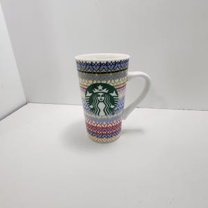 May include: A tall ceramic Starbucks mug featuring a white handle and a repeating geometric pattern in shades of blue, pink, and gold. The iconic green and white Starbucks siren logo is centered on the front of the mug against the colorful design.