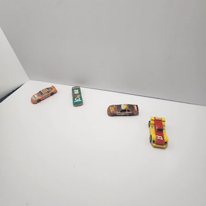 Puede incluir: Cuatro coches de carreras de juguete en naranja, verde, marrón y amarillo con detalles en rojo. Los coches tienen varios números de carreras y logotipos de patrocinadores. Sobre una superficie blanca.
