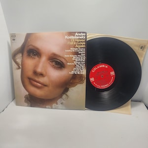 Puede incluir: Una portada del álbum "I'll Never Fall In Love Again" de Andre Kostelanetz con un retrato de mujer. El disco de vinilo es negro con una etiqueta roja y blanca. La portada del álbum enumera los títulos de las canciones.