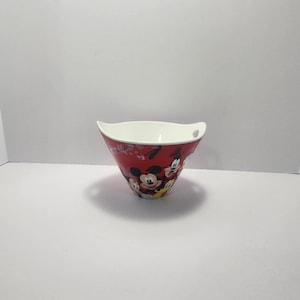Op de afbeelding: Een rode keramische kom met een witte binnenkant. De buitenkant heeft een ontwerp van Mickey Mouse, Donald Duck en Goofy. De kom heeft een unieke vorm met gebogen zijkanten en kleine handvatten. Een decoratief item voor de keuken.