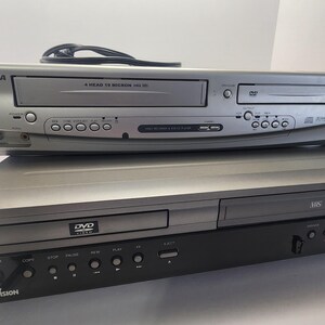 Może przedstawiać: Dwa srebrne i czarne odtwarzacze wideo są ułożone jeden na drugim. G&oacute;rny odtwarzacz to combo VHS i DVD z napisem "4 HEAD 19 MICRON HQ VHS". Dolny odtwarzacz to odtwarzacz DVD z napisem "DVD VIDEO".