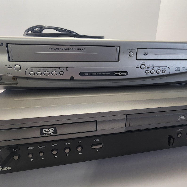 Może przedstawiać: Dwa srebrne i czarne odtwarzacze wideo są ułożone jeden na drugim. G&oacute;rny odtwarzacz to combo VHS i DVD z napisem "4 HEAD 19 MICRON HQ VHS". Dolny odtwarzacz to odtwarzacz DVD z napisem "DVD VIDEO".
