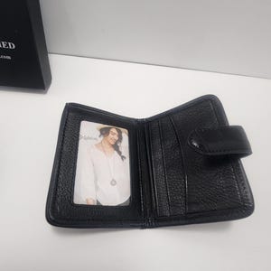 May include: Open black leather wallet with a snap closure. The wallet has multiple card slots and a clear ID window. A photo of a woman is visible in the ID window. The wallet is on a white surface.