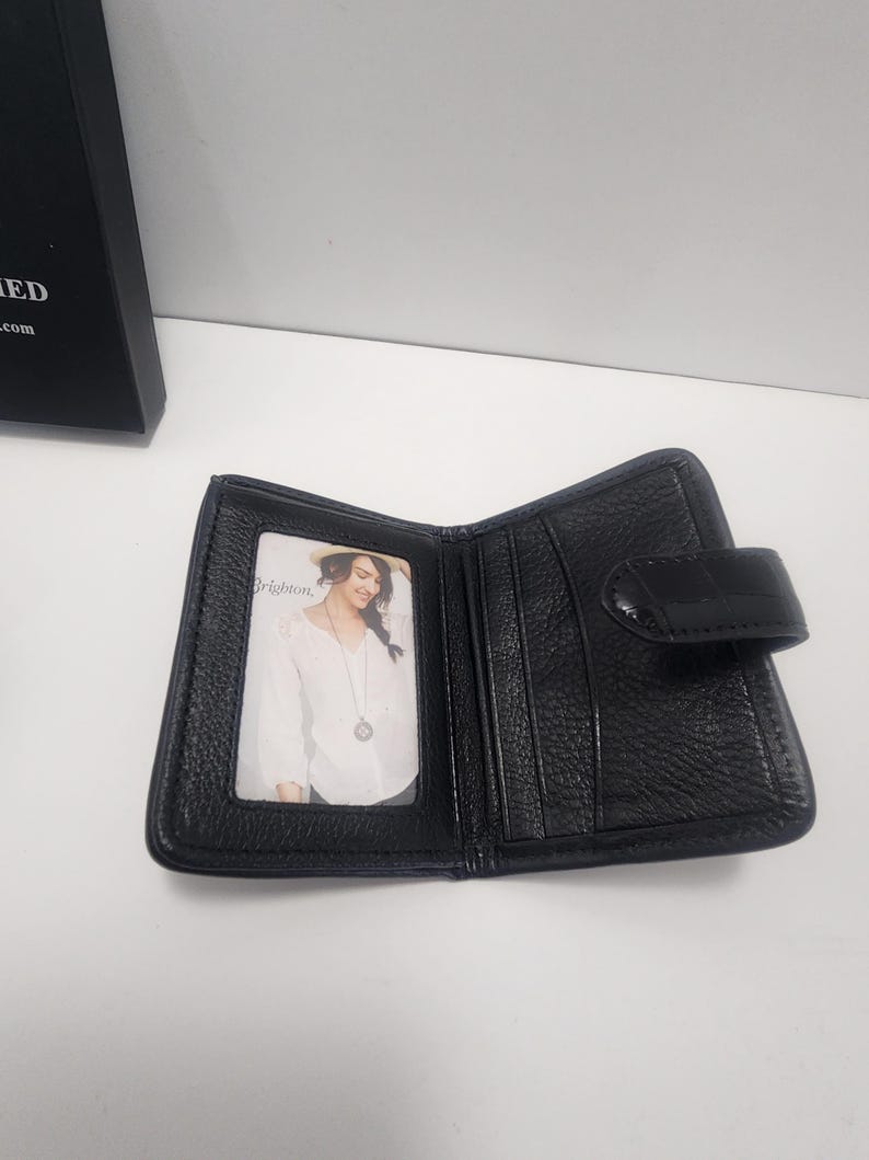 May include: Open black leather wallet with a snap closure. The wallet has multiple card slots and a clear ID window. A photo of a woman is visible in the ID window. The wallet is on a white surface.