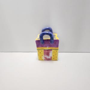 May include: A small, ceramic gift bag-shaped container. The container is painted with a yellow and purple color scheme, with red polka dots and blue handles. The container is likely used for decorative purposes.