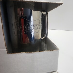 Puede incluir: Una taza de Budweiser plateada en una caja de cart&oacute;n blanca. La taza tiene un asa y el logotipo de Budweiser est&aacute; grabado en el lateral.
