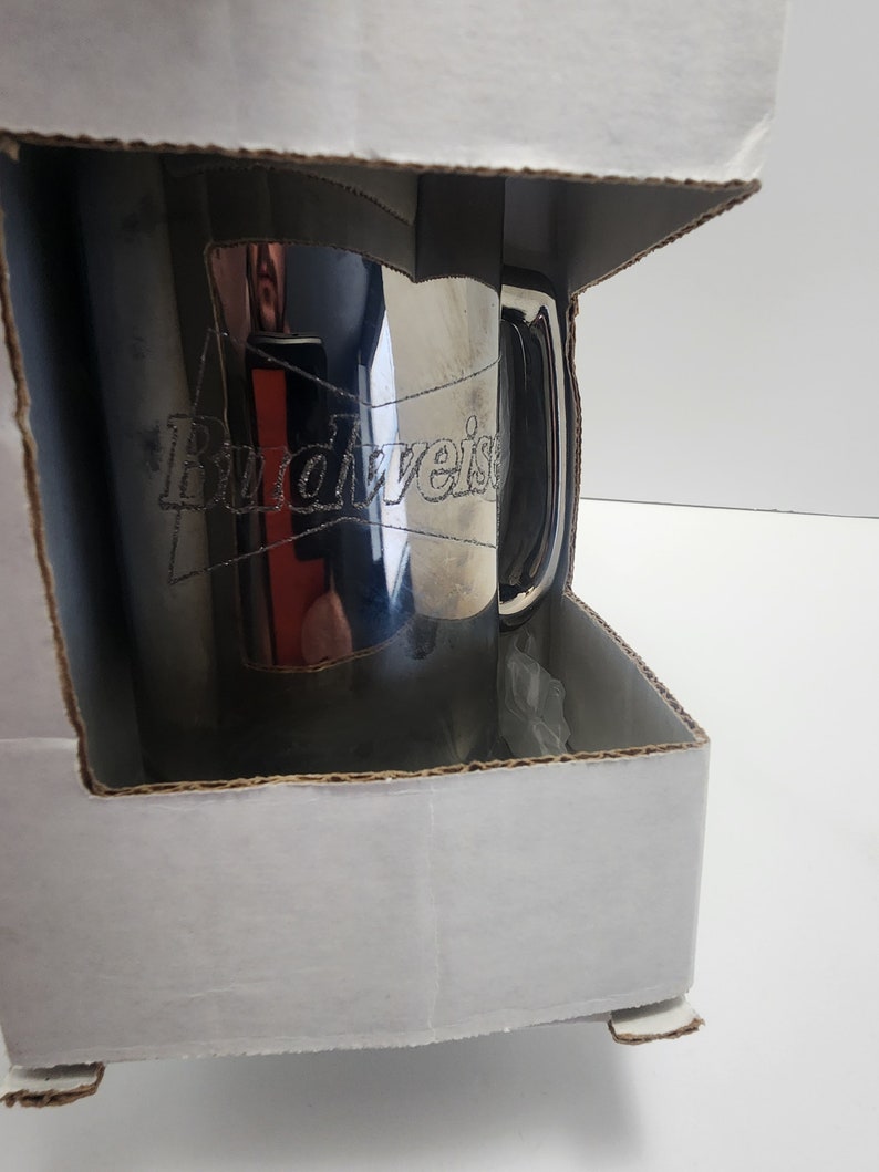 Puede incluir: Una taza de Budweiser plateada en una caja de cart&oacute;n blanca. La taza tiene un asa y el logotipo de Budweiser est&aacute; grabado en el lateral.