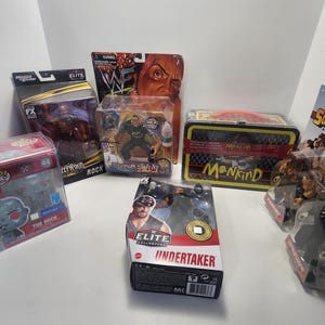 May include: A collection of wrestling action figures in their packaging. The figures include The Rock, Mankind, and The Undertaker. The packaging is colourful and features the wrestlers' names and logos.