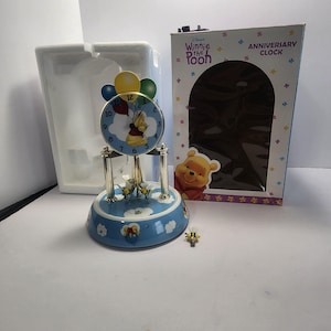 May include: A Winnie the Pooh anniversary clock with a blue base and a clock face featuring Pooh Bear holding balloons. The clock has gold-coloured accents and comes with a decorative box.