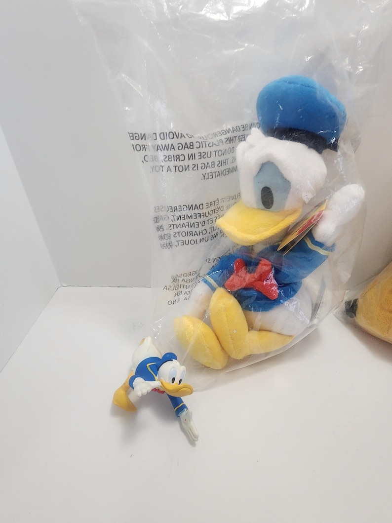 Pu&ograve; includere: Un peluche di Paperino in un sacchetto di plastica trasparente, con cappello da marinaio blu, giacca e papillon rosso. &Egrave; presente anche una piccola statuina di Paperino.